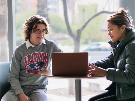 two students sitting at a table looking at a laptop