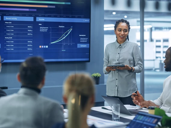 A person presenting financial information to a boardroom in front of a digital display.