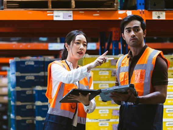 Two people wearing safety jackets and holding tablets while working in a warehouse. 