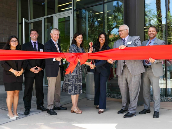 A group of people in front of a building cutting a big red ribbon with giant scissors.