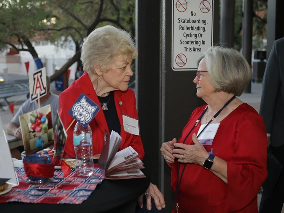 Two Norton alumni dressed in Wildcat red chat around a festive table at the Homecoming celebration