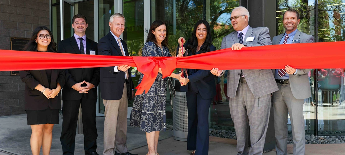 A group of people in front of a building cutting a big red ribbon with giant scissors.
