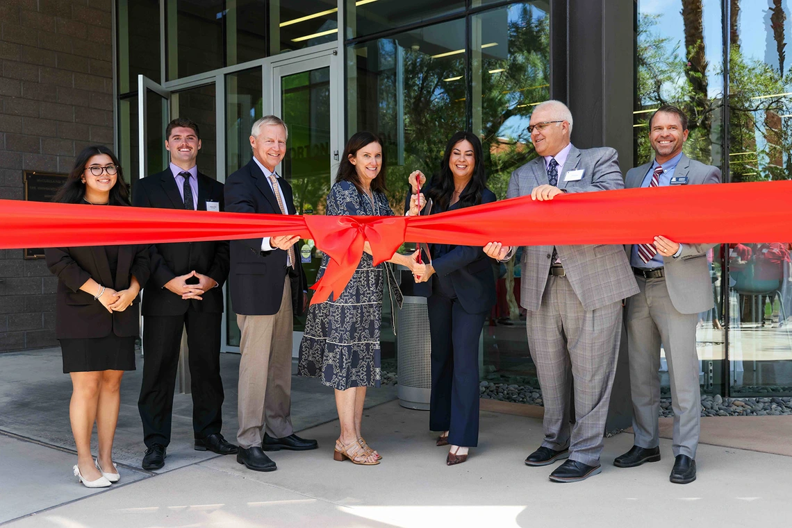 A group of people in front of a building cutting a big red ribbon with giant scissors.