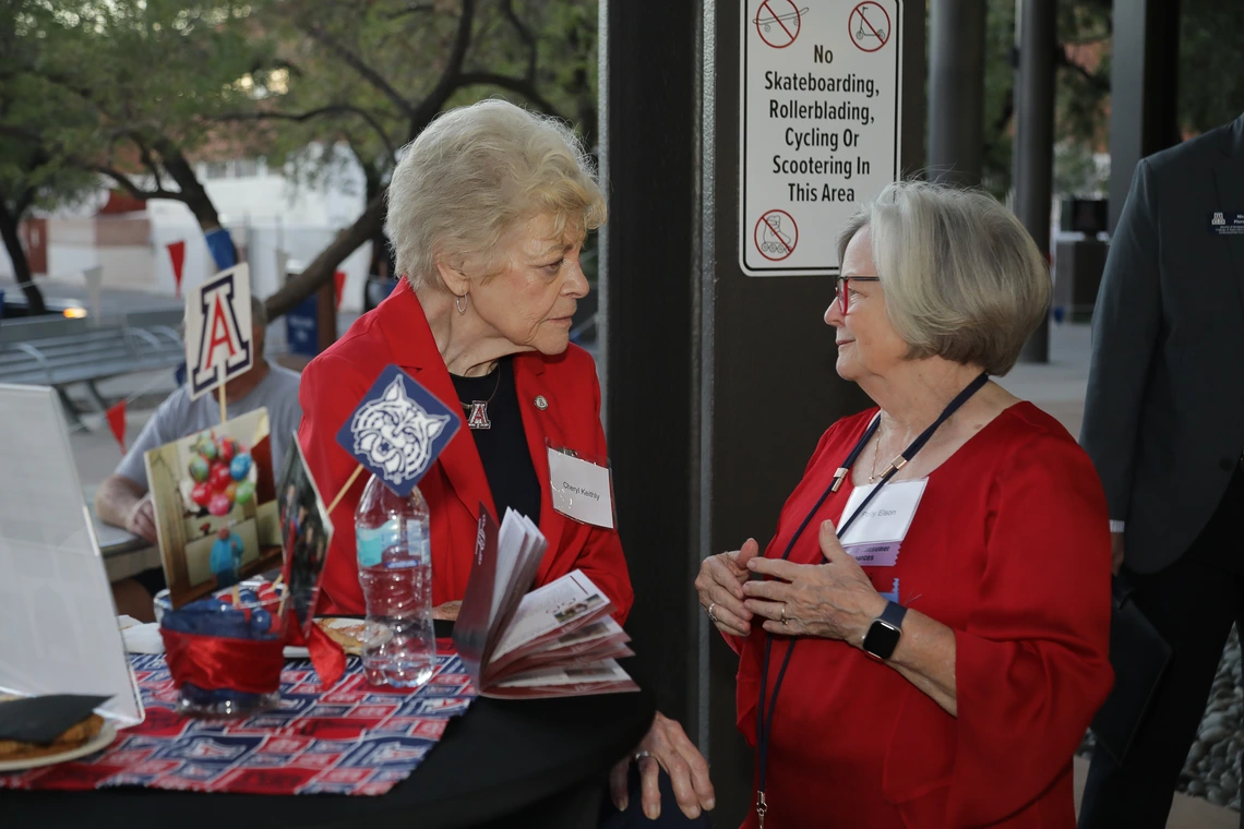 Two Norton alumni dressed in Wildcat red chat around a festive table at the Homecoming celebration
