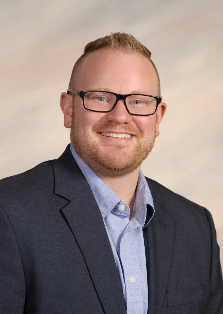 Photo of Russ; light skin with red hair, glasses, and a dark blue suit jacket and light blue button up shirt